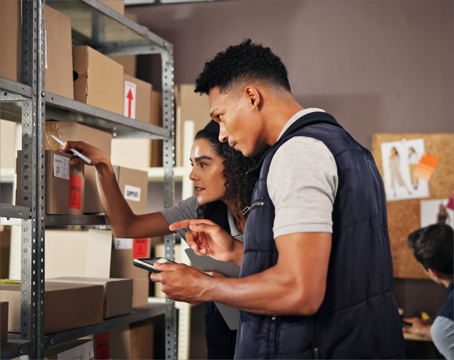 Two warehouse workers are actively scanning inventory labels on boxes on a shelf in a stockroom. The male worker is pointing at a handheld inventory management device held by the female worker, illustrating the use of mobile stock control and barcode scanning for fast and accurate order fulfillment and inventory tracking.