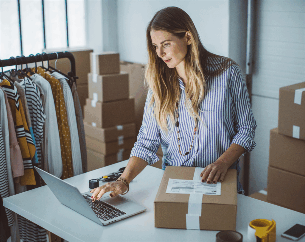 Small business owner preparing packages and updating product listings on a laptop in a home office filled with shipping boxes and clothing items.