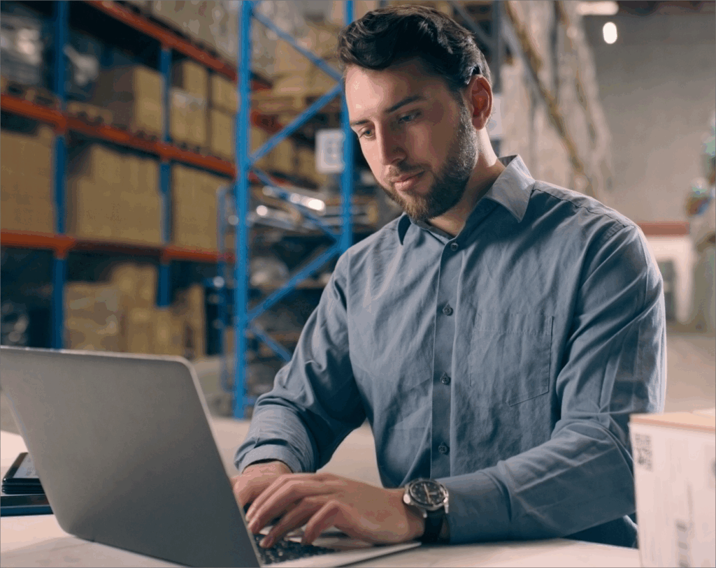 Man working on a laptop in a warehouse, analyzing data and managing inventory operations.