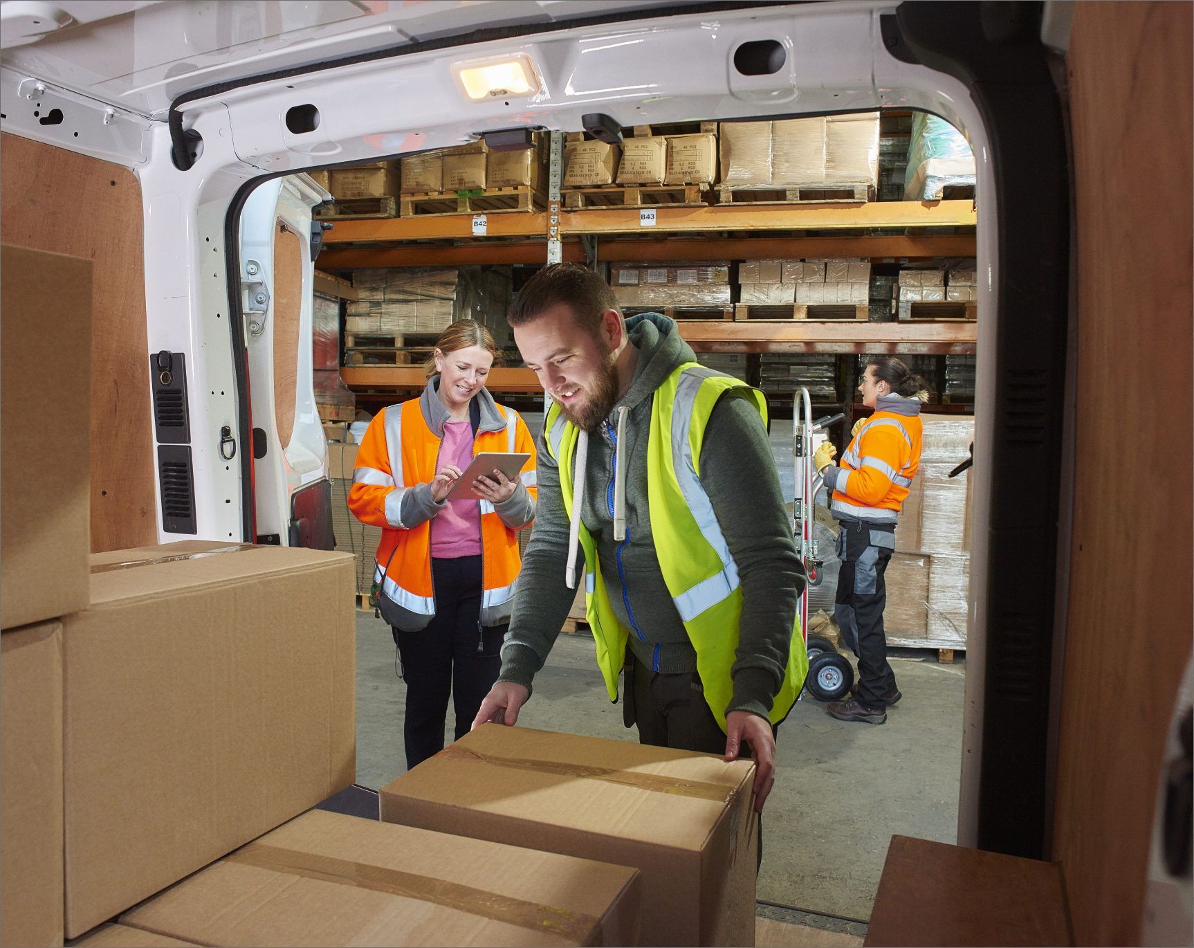 Three warehouse staff in high-visibility gear efficiently loading packages into a delivery van, with one worker using a tablet for digital dispatch. This image showcases streamlined shipping processes and outbound logistics managed by Linnworks Shipping Management. It highlights accurate package tracking, efficient van loading, and coordinated delivery operations for eCommerce order fulfillment, emphasizing speed and precision from the warehouse.