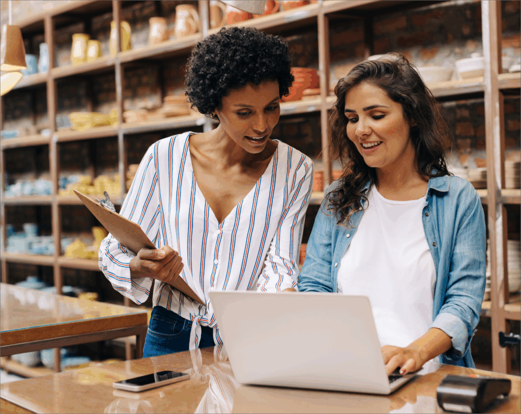 Two colleagues reviewing sales and inventory data on a laptop inside a store, discussing restocking and stock forecasting plans.