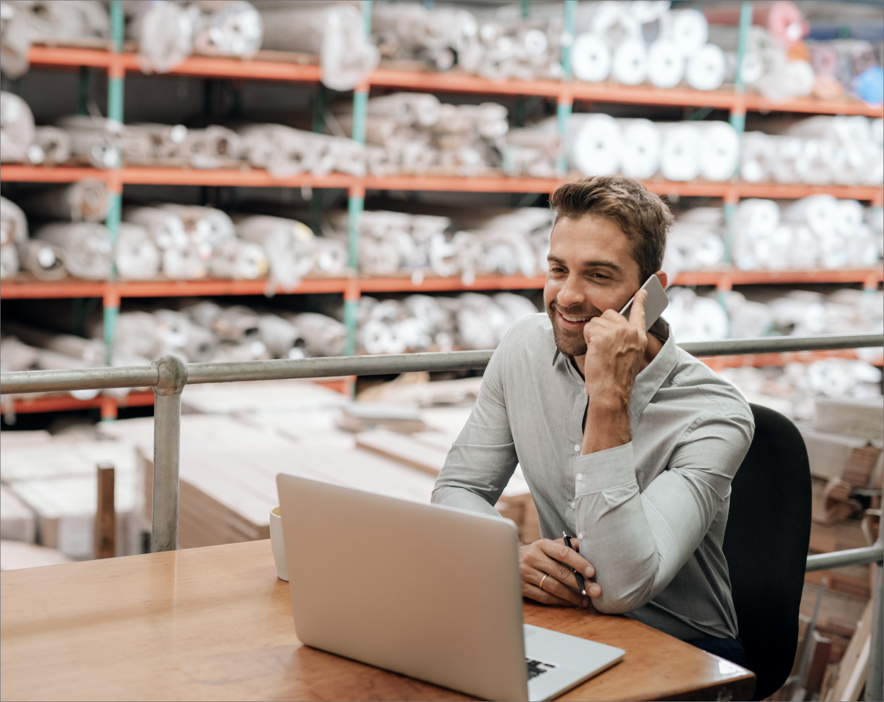 A smiling warehouse manager or business owner on a phone call, working on a laptop in a well-organized warehouse environment. This image represents the crucial communication and remote warehouse management possible with Linnworks WMS, enabling efficient inventory control, supply chain coordination, and order fulfillment from any location.