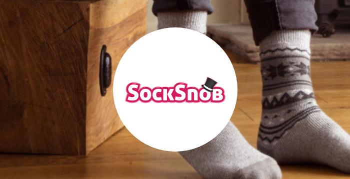 Person wearing patterned grey socks beside a wooden chest, with the SockSnob logo displayed in the center.